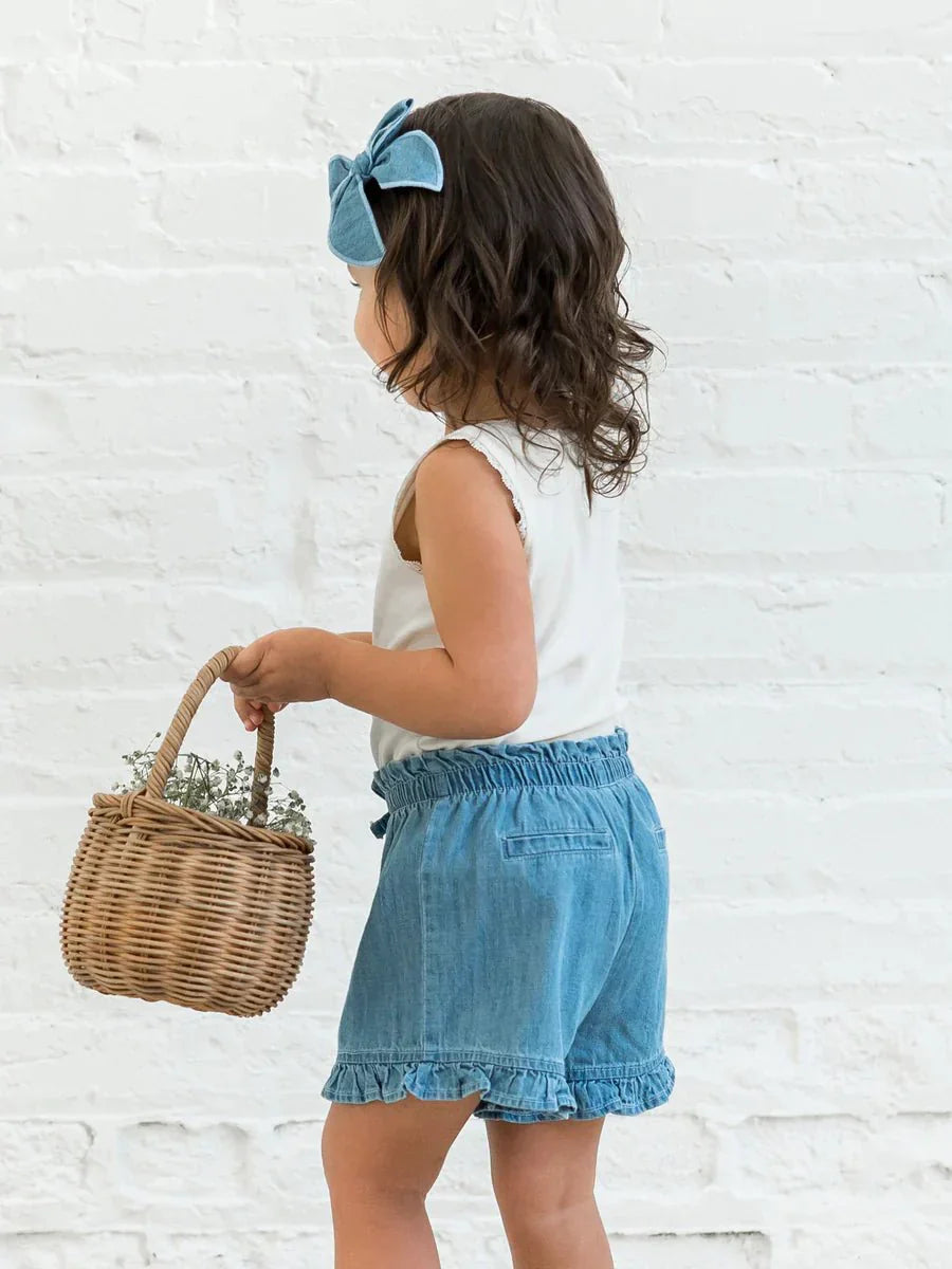 Little girl wearing Callie Chambray Ruffle Waist Shorts with a basket against a white brick wall.