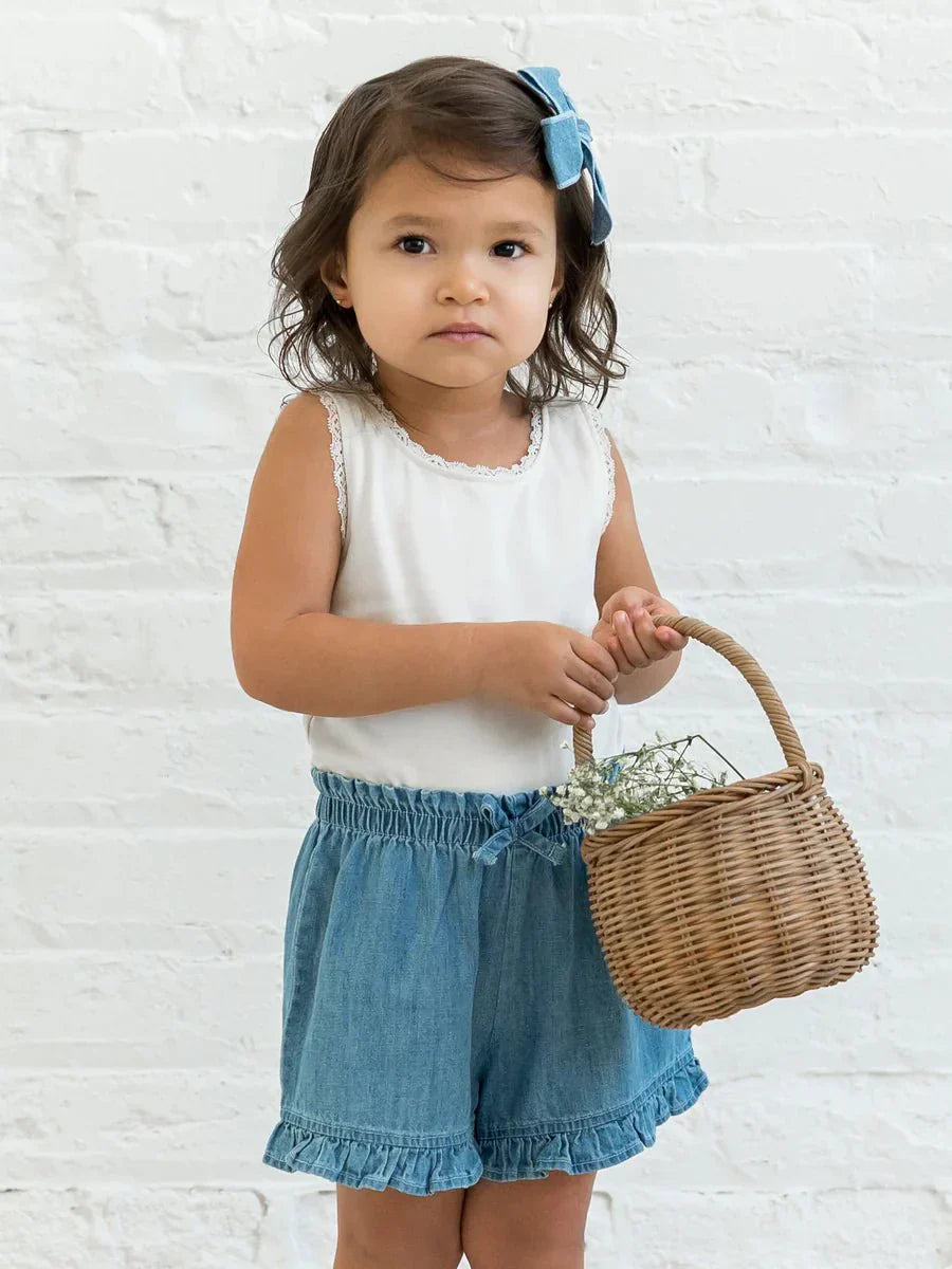 Child wearing Callie Chambray Ruffle Waist Short with basket, white background.